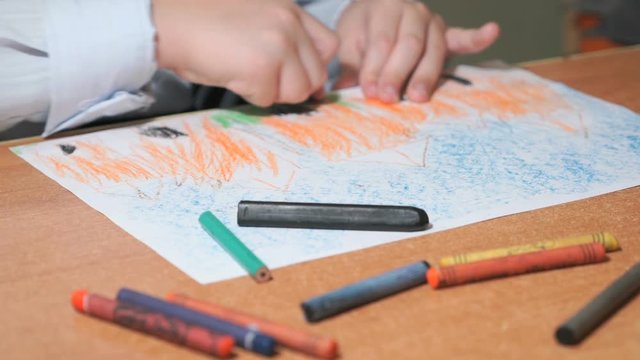 Close-up of unknown little child dressed in shirt with tie draws the pictures using color chalks and pencils in the album in the kindergarten