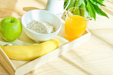 Wooden Tray with Healthy Breakfast Ingredients on Table on Terrace. Oats in Bowl Nut Milk in Pitcher Orange Juice Banana Green Apple. Bright Morning Sunlight. Healthy Food. Copy Space. Tilted