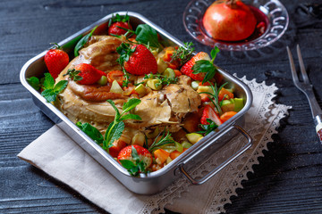 Whole wild rabbit backed in pomegranate juice with strawberry, vegetables, spices and herbs in metallic dish on old black rustic wooden background. Clean eating, healthy food and diet concept