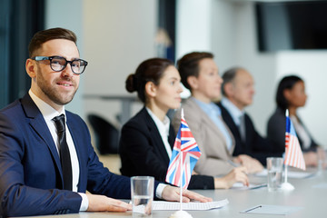Elegant delegate in suit and eyeglasses looking at camera at political conference on background of other participants
