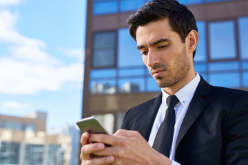 Young employee in suit and tie reading or writing message in smartphone in urban environment