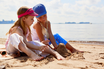 Two barefoot girls sitting on beach and building tower from sand by seaside on summer day