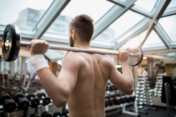 Back view of shirtless man keeping heavy barbell on his shoulders while weightlifting in gym