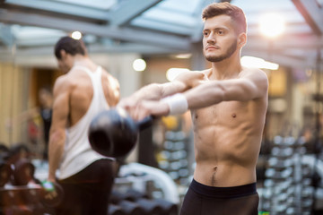 Fototapeta na wymiar Shirtless guy with stretched arms lifting heavy kettlebell while exercising in modern sports club
