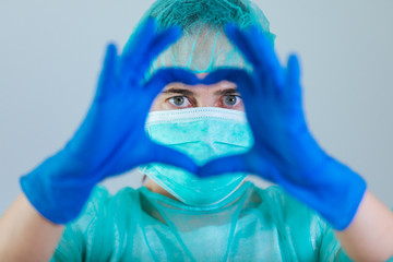 Young woman doctor with blue eyes wearing a cap and face mask showing hands in shape of love heart in front of face with gloves