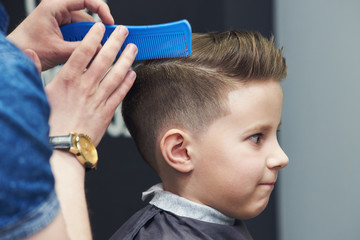 Barber is using styling gel and hairbrush to make a styling to a Caucasian boy.