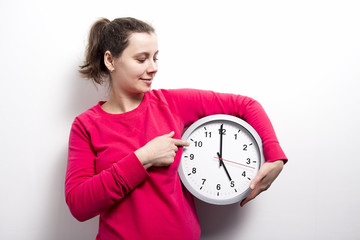 brunette girl is holding clock and points to hands of clock on white background. concept of time. Watch, stop and control time. Young woman holds round clock in hands.