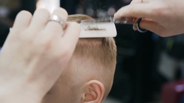 Close-up. Professional hairdresser makes a new haircut to the little boy. During haircuts barber uses an scissors and a comb. The guy's haircut into a man's hairdressing salon,