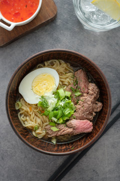 Asian ramen soup with beef, egg, chives in bowl on grey background.