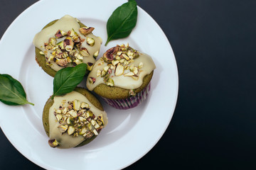 Green basil muffins with white chocolate frosting on the white plate with black background. Basil leaves and pistachio topping. Healthy sweets variation. Dark food photography overhead composition