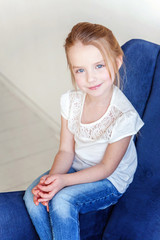 Fototapeta premium Sweet little girl in jeans and white T-shirt at home sitting on modern cozy blue chair relaxing in white living room
