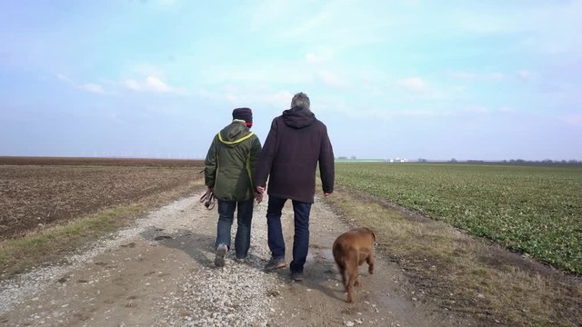 Senioren Paar mit Hund gehen Gassi, Mann und Frau laufen nebeneinander auf dem Feldweg
