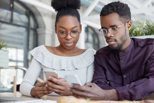Horizontal shot of dark skinned young female and male freelancers type text notifications on smart phones, chat online, meet together for making common work, watch video from social networks