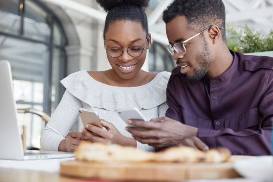 Happy African American couple of enterpreneurs develop new business strategy on portable laptop computer, use mobile phones for surfing information in internet, sit together in spacious office room