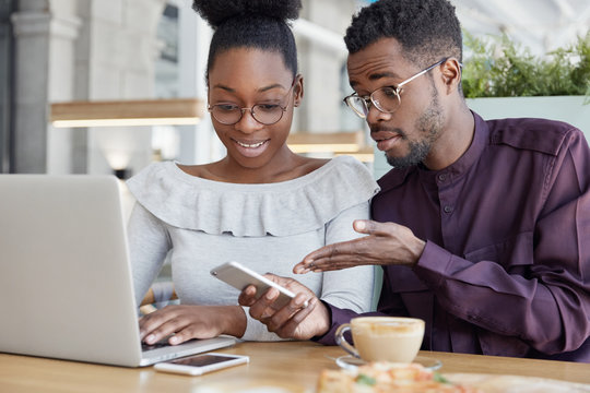 Two dark skinned male, female administrative managers check messages on cellular, keyboard on laptop computer, check information, have problems with contact of service operator, sit in coworking space