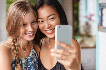 Fototapeta premium Happy two mixed race women smile and pose in front of camera, use modern cell phone for making photos, hug and laugh happily, sit against cafe interior. Asian female meets with friend, take selfie