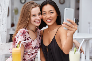 Cheerful two mixed race female friends, make photo in front of smart phone, recreat together in cafeteria, drink cocktails, pose for selfie. Glad Asian woman takes pictures for publication online