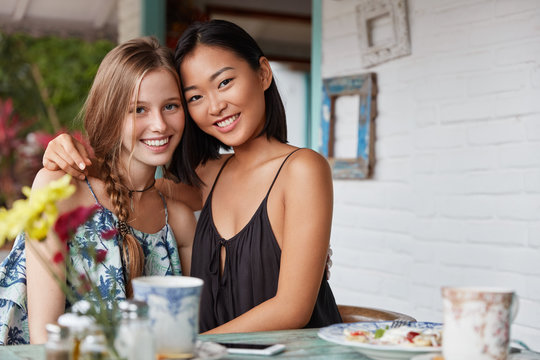 Mixed race beautiful young women of different nationalities embrace as demonstate interracial true friendship or relationships, drink coffee in bar, meet after long departure, miss each other