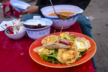 Street food, consisting of rice pancakes with quails eggs, pork and beef, on a bed of green leaves, has a honey and chilli sauce poured over it, in Hoi An, Vietnam