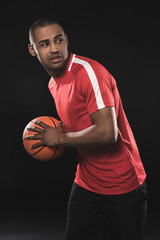 Full length portrait of handsome guy playing basketball. He looking carefully to the side and hiding ball over his body. Isolated on black background