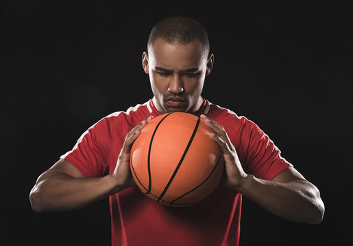 Waist up of handsome sportsman in red t-shirt. He squeezing the basketball two hands in front of the chest while hard watching at him. Isolated on black background