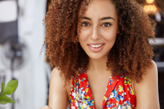 Close up shot of beautiful dark skinned curly woman looks with pleased expression, comes on meeting with colleague to discuss collaboration, pose at cafe interior. Positive African American female