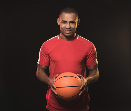 Waist up portrait of handsome guy keeping basketball in red t-shirt. He looking at camera and smiling happily. Isolated on black background