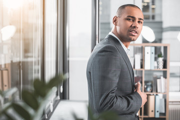 Waist up portrait of young guy standing at cabinet near window. He looking mysteriously at camera. Copy space in left side