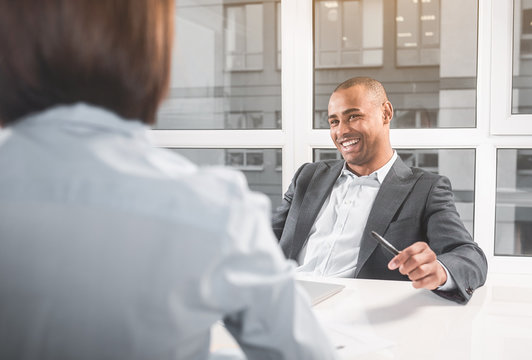 Young african man enthusiastically speaking with woman. He keeping pen while looking at her and playfully laughing. Focus on guy from close up of the back of girl