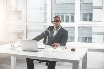 Waist up portrait of young african businessman sitting in office at desk near laptop. Man is looking calm at camera while document and glasses with cup of coffee are on table