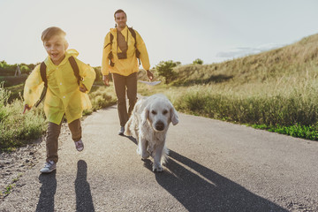 young cheerful boy with rucksack running along sunlit rode with his dog. His father walking on background. Copy space in right side. Full length portrait - Powered by Adobe