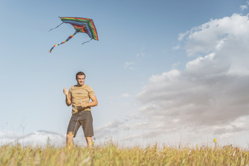 Glad adult man standing on sunlit field, colorful kite flying in air above him. Copy space in right side