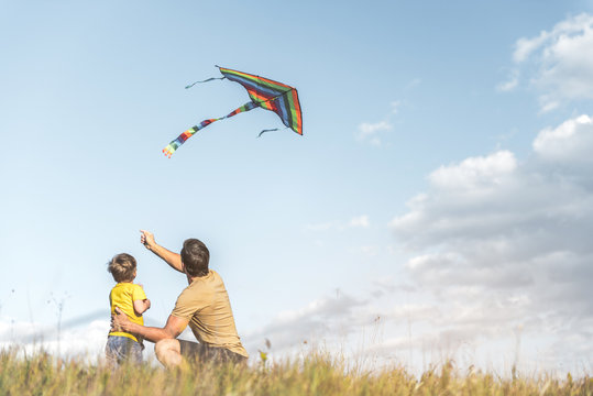 Parent And Little Boy Are Sitting With Their Backs To Camera On Field And Flying Kite. Copy Space In Right Side