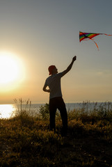 silhouette of active young man in cap launching colorful kite flying in air at summer sunset near sea