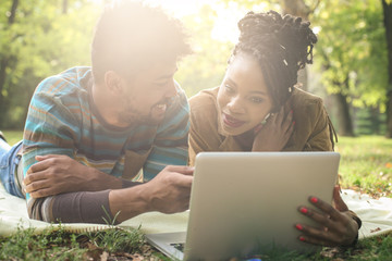 Cheerful African American couple lying down in park and using laptop.