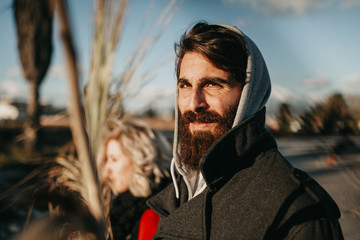 Hipster couple portrait with focus in the man, looking at camera. Cool and weird young couple posing outdoors while holding palm tree branches. - obrazy, fototapety, plakaty