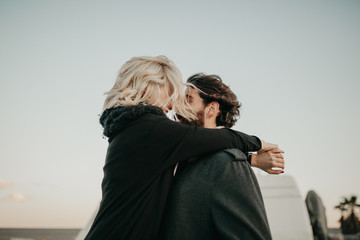 Cool young couple kissing each other outdoors while they are embraced, during a road trip stop, with their van in the background.