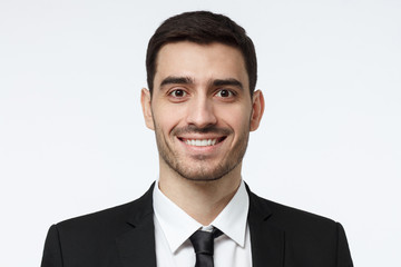 Closeup headshot of business man standing against grey background smiling with satisfaction and confidence
