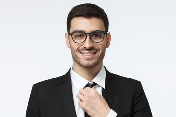 Closeup shot of young business man in spectacles pictured isolated on grey background, straightening his tie, looking positive and confident, satisfied with success of his team