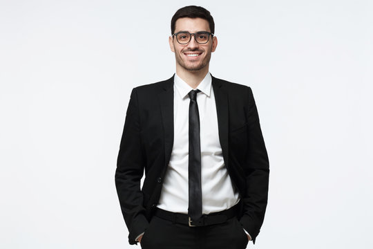 Portrait of a cheerful business man dressed in black suit, posing  standing with hands in pockets and looking at camera isolated on grey background
