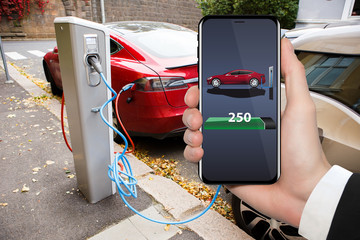 Hand with phone on a background of electric car charging point. On a device screen indicator of...