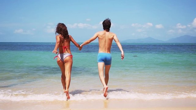 Young slim couple holding hands running into tropical sea on sunny day. slow motion. 1920x1080