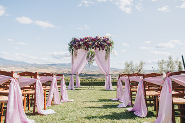 arch, decorated with trunks and flowers, stands in the wedding ceremony area on the hillside in Tuscany, as are the chairs for guests
