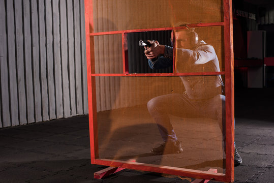 An adult man in a denim shirt shoots a gun in a shooting range through a red obstacle