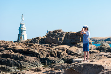 The boy in the striped T-shirt and a cap on the rock. In the background stands a lighthouse.