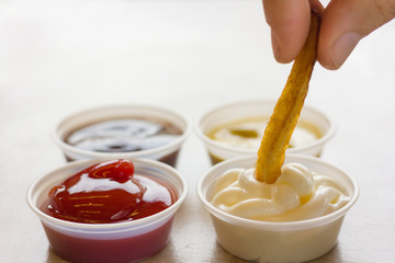 Close up on french fries dipping in mayonnaise with fingers next to plastic dips of ketchup...