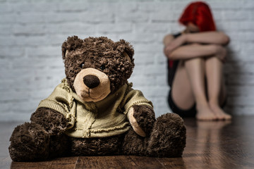 In the foreground is a soft toy-a bear. Behind the toy of a girl sitting near brick wall.
