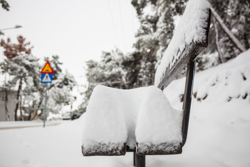 Snow covers a wooden bench. Misty sky and blurred snowy trees background. Close up view.