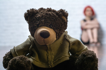 In the foreground is a soft toy-a bear. Behind the toy of a girl sitting near brick wall.
