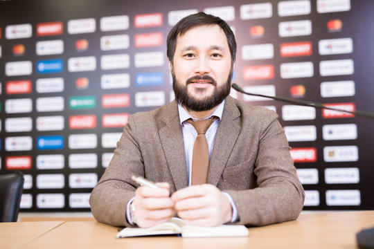 Waist-up portrait of handsome bearded entrepreneur looking at camera while sitting against brand wall and participating in press conference